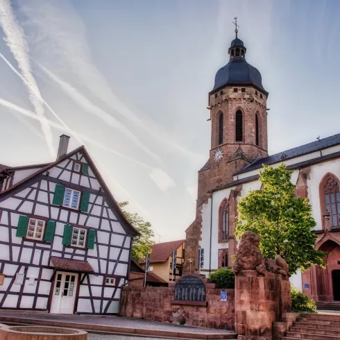 Eine malerische Altstadt mit einem Fachwerkhaus und einer imposanten Kirche. Der Himmel ist klar und es gibt sichtbare Flugstreifen.