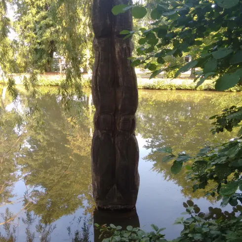 Eine Holzskulptur ragt aus einem ruhigen Teich. Umgeben von Pflanzen und Bäumen reflektiert das Wasser das Licht.