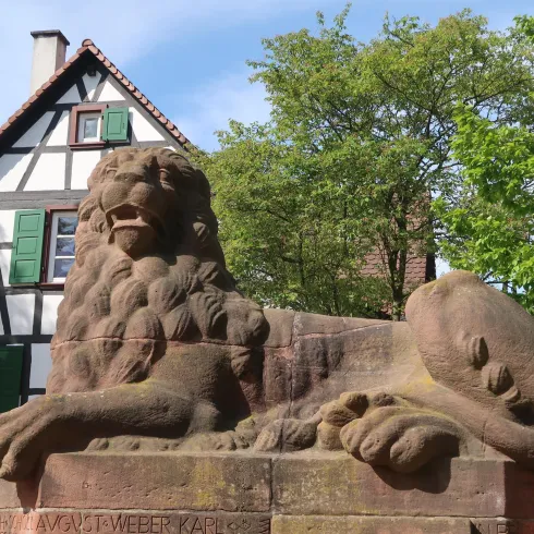 Ein steinernene Löwe sitzt vor einem historischen Fachwerkhaus. Im Hintergrund sind Bäume und ein blauer Himmel zu sehen.