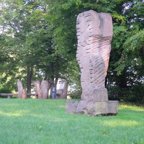 Eine große Steinfigur steht in einem grünen Park. Im Hintergrund sind weitere Kunstwerke und Bäume sichtbar.