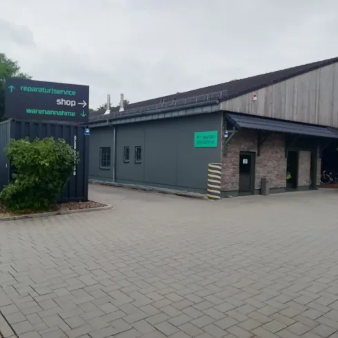 Ein modernes Gebäude mit einer grauen Fassade und Holzverkleidung. Vor dem Eingang befindet sich ein gepflasterter Bereich und einige Bäume.