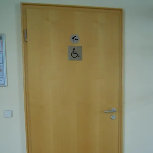 Eine Holztür mit einem Schild für die behindertengerechte Toilette. Es gibt Symbole für Männer und die Barrierefreiheit.