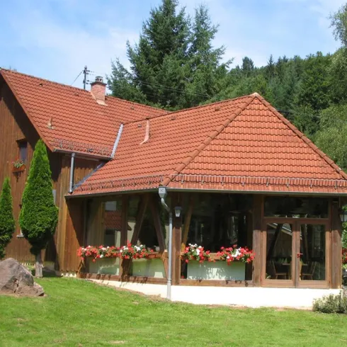 Ein schönes Holzhaus mit rotem Dach und bunten Blumen vor den Fenstern. Umgeben von grünen Bäumen und einer gepflegten Wiese.