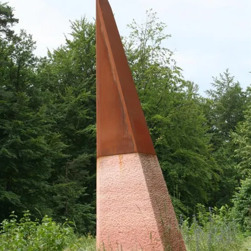 Ein hoher, spitzer Kunststein steht inmitten von Bäumen. Der untere Teil hat eine helle Farbe, während die Spitze rostig aussieht.