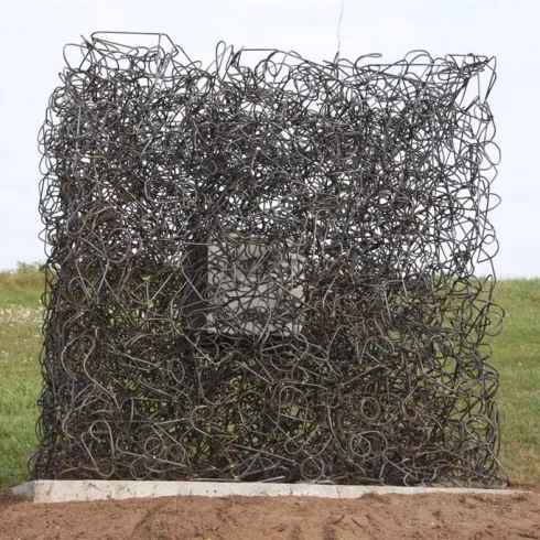 Eine künstlerische Skulptur aus verworrenem Draht bildet einen quadratischen Raum. Sie steht auf einem offenen Feld mit grünem Gras im Hintergrund.