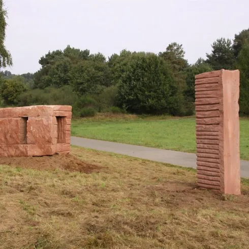 Eine interessante Skulptur aus Stein, die in einer grünen Landschaft steht. Im Hintergrund sind Bäume und ein Weg sichtbar.