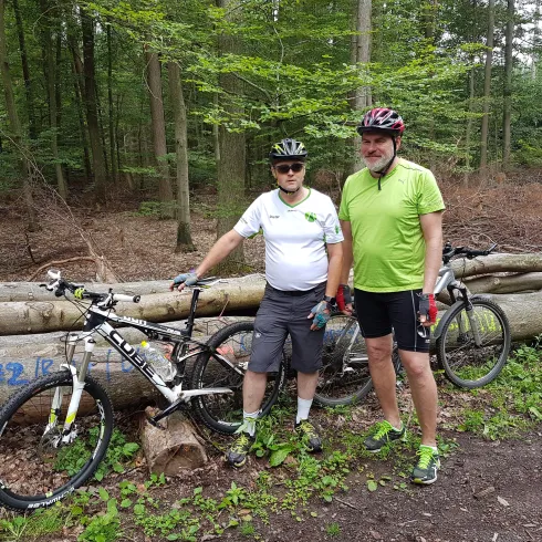 Zwei Männer stehen neben ihren Mountainbikes im Wald. Sie tragen Fahrradhelme und sportliche Kleidung.