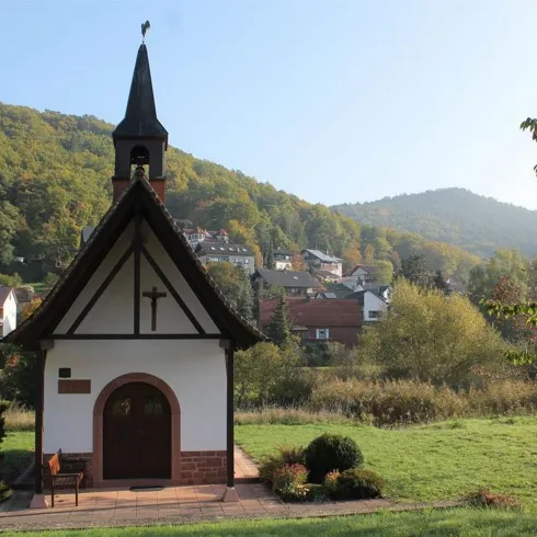 Eine kleine Kapelle umgeben von grünen Wiesen und Hügeln. Im Hintergrund sind malerische Häuser in einer ruhigen Landschaft zu sehen.