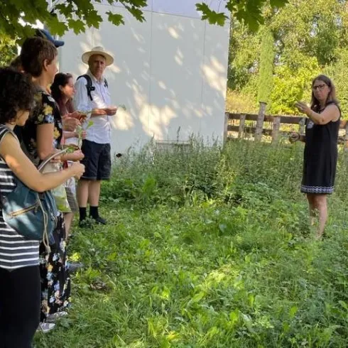Eine Gruppe von fünf Personen steht in einem grünen Garten. Eine Frau erklärt etwas, während die anderen aufmerksam zuzuhören.