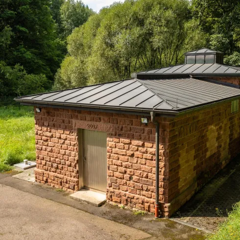 Ein modernes Gebäude aus roten Steinen in einer grünen Umgebung. Es hat ein flaches, schwarzes Dach und ist von Bäumen umgeben.