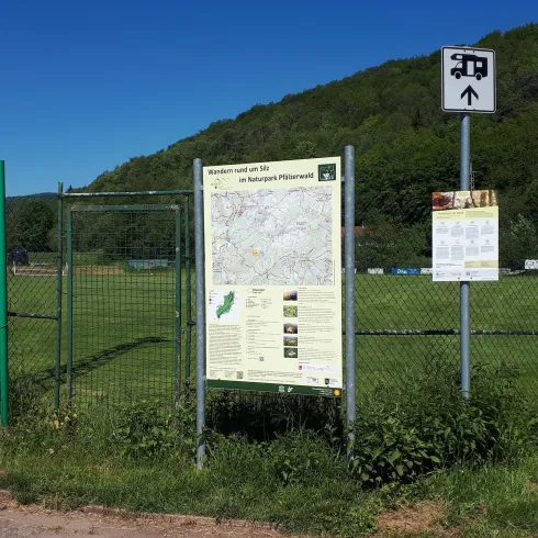 Ein Informationsschild und eine Karte auf einem Sportgelände. Im Hintergrund sind Bäume und ein klarer Himmel sichtbar.
