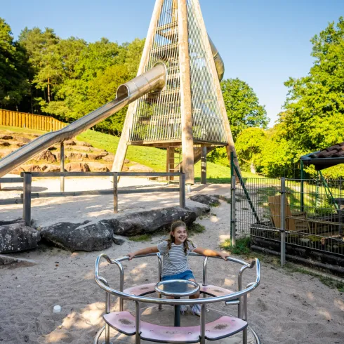 Ein Kind spielt auf einem Karussell in einem Spielplatz. Im Hintergrund steht eine große Rutsche unter klarem Himmel.