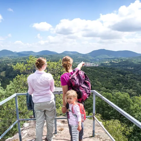 Drei Wanderer stehen auf einer Aussichtsplattform und betrachten die Landschaft. Im Hintergrund sind grüne Hügel und ein blauer Himmel mit Wolken zu sehen.