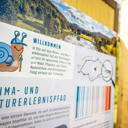 Ein Willkommensschild mit einer Zeichnung einer Schnecke und Informationen über den Klima- und Naturerlebnispfad. Es gibt Hinweise zur Erkundung von Wald, Pflanzen und Tieren in der Umgebung.