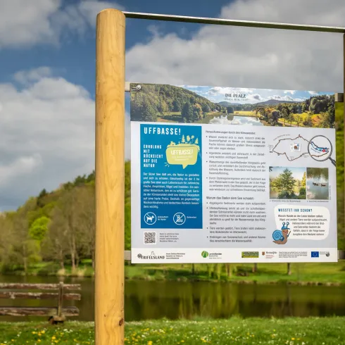 Ein Informationsschild am Ufer eines ruhigen Gewässers. Im Hintergrund sind Bäume und ein blauer Himmel mit wenigen Wolken zu sehen.