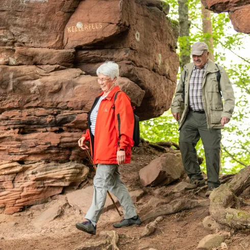 Ein älteres Paar wandert auf einem schmalen Pfad zwischen großen Felsen. Die Frau trägt eine rote Jacke und der Mann eine helle Kapuzenjacke.