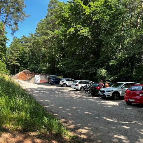 Ein Parkplatz im Wald mit mehreren Autos. Im Hintergrund sind grüne Bäume und ein blauer Himmel zu sehen.