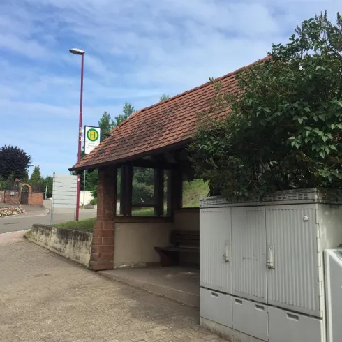 Eine Haltestelle mit einem überdachten Wartenbereich und angrenzenden Briefkästen. Im Hintergrund sind Bäume und ein klarer Himmel zu sehen.