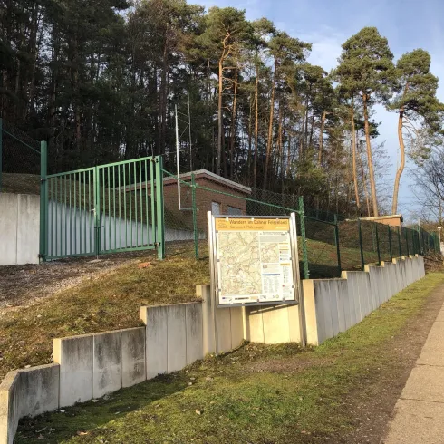 Ein Zugang zu einem abgegrenzten Bereich, umgeben von Bäumen. Ein Orientierungsschild steht an der Seite des Wegs.