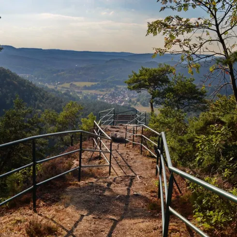Ein Wanderweg mit einem Geländer führt zu einem malerischen Blick auf das Tal. Die umgebende Natur ist grün und bewaldet, und der Himmel ist leicht bewölkt.