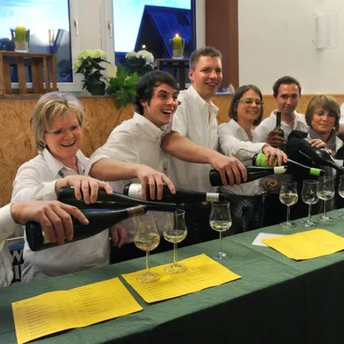 Eine Gruppe von sechs Personen in weißer Kleidung steht hinter einem Tisch und schenkt Wein in Gläser ein. Im Hintergrund sind einige Pflanzen und Fenster sichtbar.