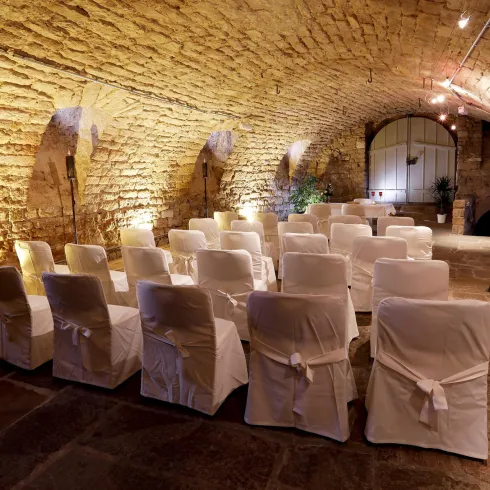 Ein stimmungsvoller, beleuchteter Kellerraum mit mehreren weißen Stühlen. Die Wände sind aus Stein, was eine romantische Atmosphäre schafft.