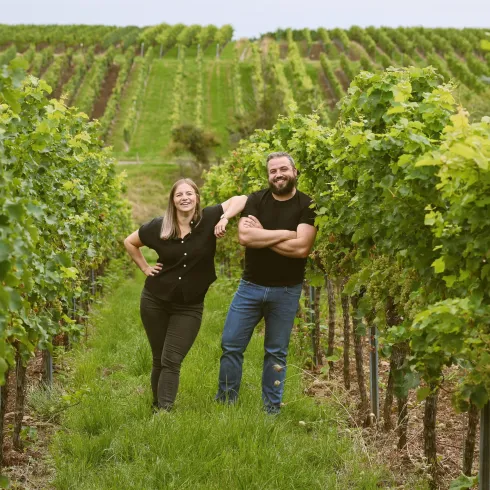 Ein Paar steht glücklich zwischen Weinreben in einem Weinberg. Im Hintergrund sind sanfte Hügel und weitere Rebstöcke zu sehen.
