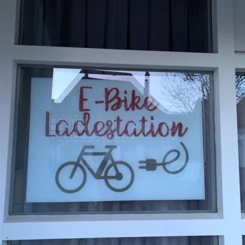 Ein Fenster mit einem Schild, das „E-Bike Ladestation“ zeigt. Darunter ist ein stilisiertes E-Bike und ein Ladekabel abgebildet.