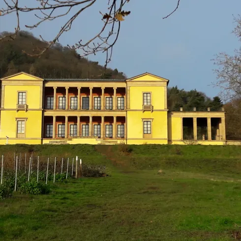 Ein gelbes historisches Gebäude mit Säulen und großen Fenstern. Es liegt in einer grünen Umgebung mit Bäumen im Hintergrund.