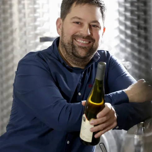 Ein lächelnder Mann hält eine Weinflasche in der Hand. Im Hintergrund sind silberne Tanks zu sehen.