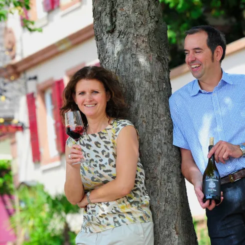 Ein Paar steht entspannt vor einem Gebäude. Die Frau hält ein Glas Wein, während der Mann eine Flasche Wein zeigt.