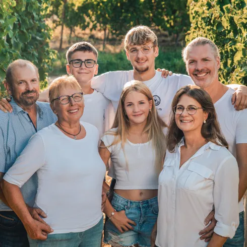 Eine Gruppe von sechs Personen steht zwischen Weinreben. Sie lächeln und tragen helle Kleidung.
