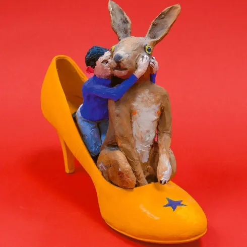 Ein gelber High Heel mit einer Figuren-Szene darin. Ein Mensch umarmt einen Hasen, während sie auf dem Schuh sitzen.