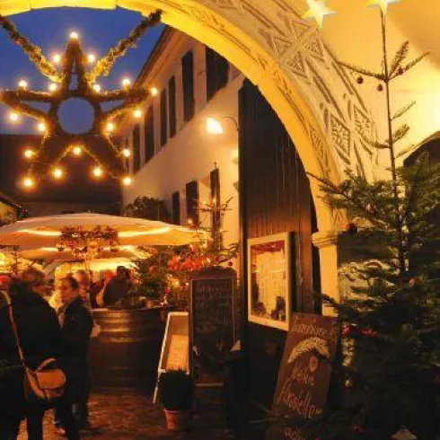Eine festlich dekorierte Gasse mit Lichtern und Tannenbäumen in der Dämmerung. Besucher genießen die weihnachtliche Stimmung an einem Verkaufsstand.