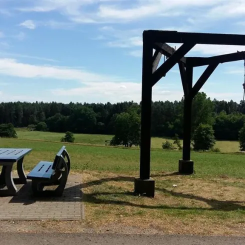 Ein Picknickbereich mit einem Tisch und Bänken steht in einer grünen Wiese. Im Hintergrund sind Bäume und ein klarer Himmel zu sehen.