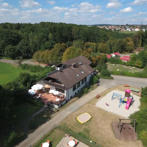 Ein Blick auf ein gemütliches Gebäude mit einer Terrasse und einem Spielplatz daneben. Umgeben von grüner Natur und einem klaren Himmel.