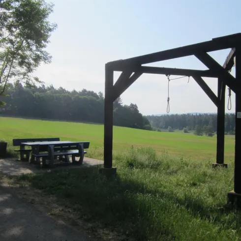 Ein großer Holzrahmen steht auf einer Wiese, umgeben von Bäumen. Neben dem Rahmen befindet sich ein Tisch mit Bänken.