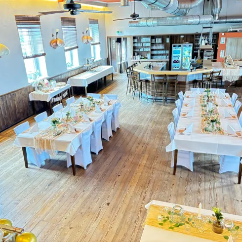 Ein helles Restaurant mit Holzboden und elegant gedeckten Tischen. Im Hintergrund befindet sich eine Bar mit Stühlen und weiteren Sitzbereichen.