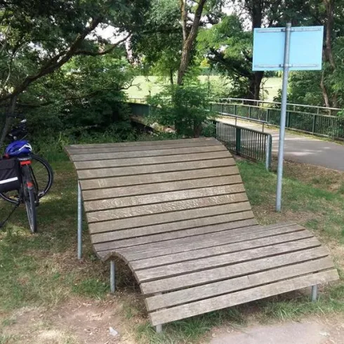 Eine moderne Holzbank steht in einem grünen Park. Im Hintergrund sind Fahrräder und Informationsschilder zu sehen.