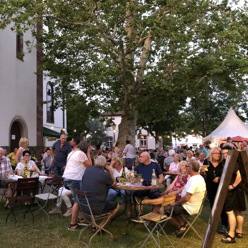 Eine lebhafte Veranstaltung im Freien mit vielen Menschen, die an runden Tischen sitzen. Im Hintergrund sind Bäume und ein Zeltdach zu sehen.