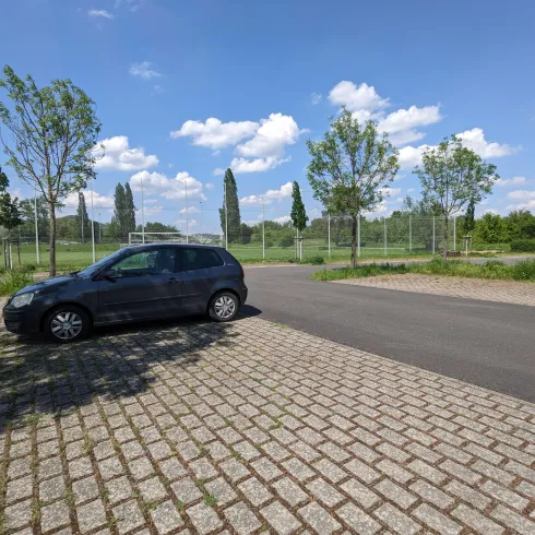 Ein Parkplatz mit einem schwarzen Auto und grünen Bäumen. Im Hintergrund sind Sportfelder und ein blauer Himmel mit einigen Wolken zu sehen.