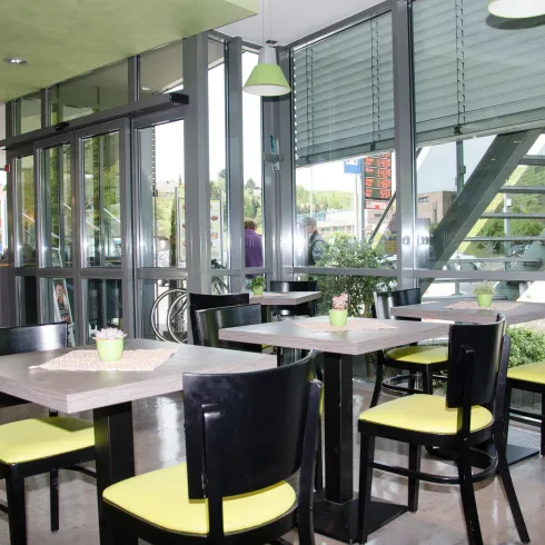 Ein modernes Café mit großen Fenstern und viel natürlichem Licht. Die Tische sind schlicht, mit schwarzen Stühlen und gelben Polstern dekoriert.