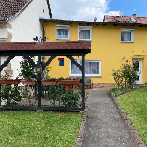 Ein gemütliches Haus mit gelber Fassade und einem kleinen Garten. Vor dem Haus steht ein pavillonähnlicher Bereich mit Rosen und Pflanzen.