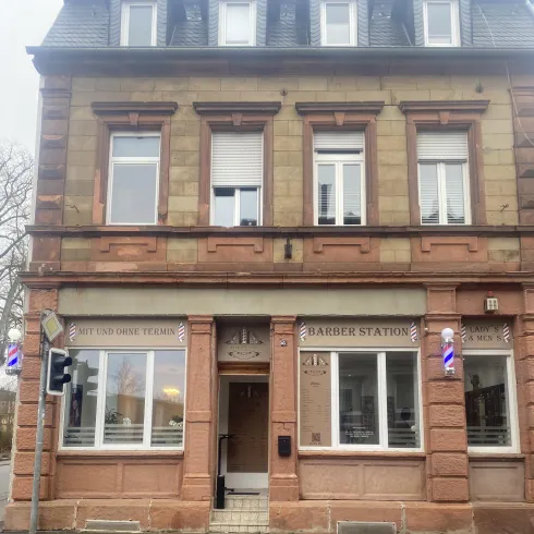 Ein traditionelles Gebäude mit einer auffälligen Fassade in rot und beige. Zwei Fenster sind sichtbar und im Erdgeschoss befindet sich ein Friseursalon.