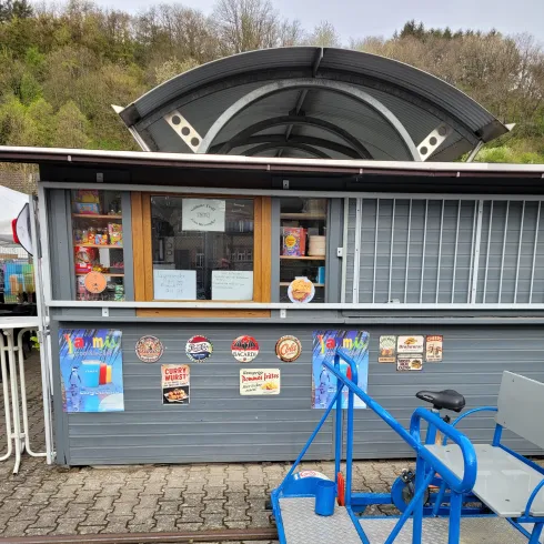 Ein Imbissstand mit einer großen Fensterfront und verschiedenen Speisekarten. Vor dem Stand steht ein blauer Anhänger.