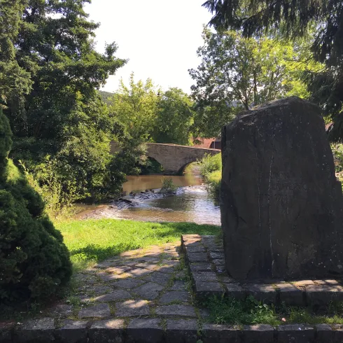 Ein ruhiger Fluss umgeben von Bäumen und dichten Sträuchern. Im Hintergrund ist eine kleine Steinbrücke zu sehen.
