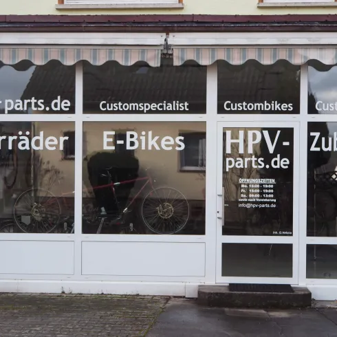 Ein Fahrradgeschäft mit großen Fenstern, das Fahrräder und E-Bikes anbietet. Die Fenster sind mit dem Namen des Geschäfts und verschiedenen Dienstleistungen beschriftet.