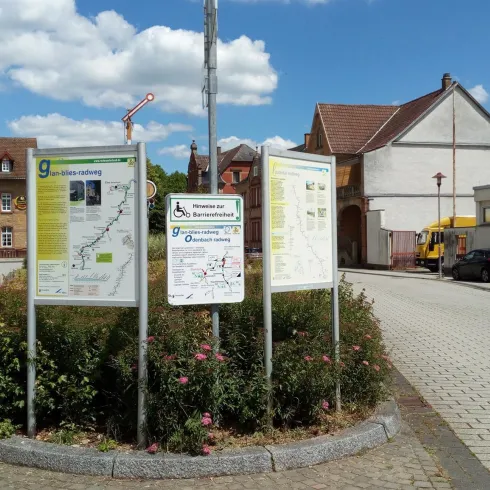 Ein öffentlicher Platz mit mehreren Orientierungstafeln und einem gepflasterten Boden. Im Hintergrund sind einige Häuser und blauer Himmel mit ein paar Wolken sichtbar.
