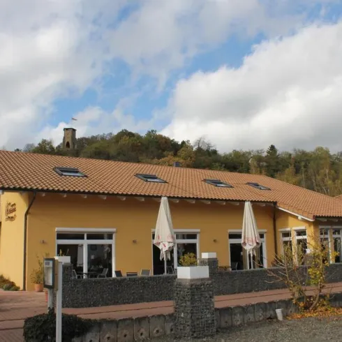Ein modernes Gebäude mit einem gelben Anstrich und einem Satteldach. Es gibt eine Terrasse mit Sonnenschirmen und eine grüne Bepflanzung im Vordergrund.