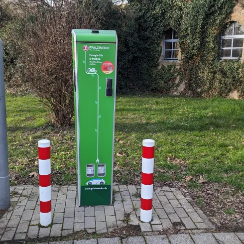 Eine grüne Ladestation für Elektrofahrzeuge steht auf einem Gehweg, umgeben von roten und weißen Pollern. Im Hintergrund sind Pflanzen und ein Gebäude sichtbar.